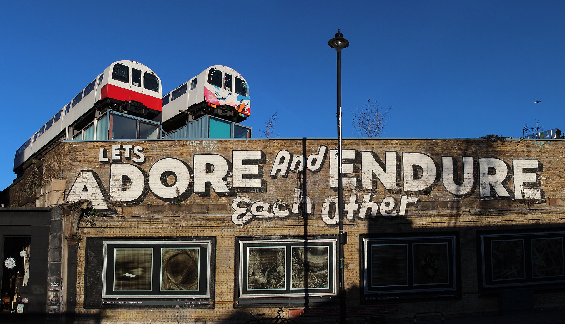 Italian Neorealism | Movies Fan Club A brick building features street art with the message 'LETS ADORE AND ENDURE' above two train cars, illustrating a scene.