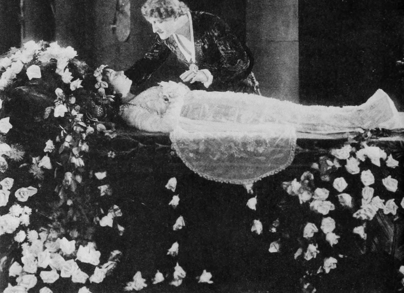A Victorian-era scene depicts mourning and death with a person grieving over a coffin surrounded by roses, reflecting.